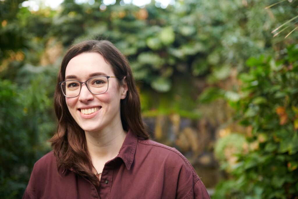 Katharina Stoll, eine junge, weiße Frau mit schulterlangen braunen Haaren und Brille steht lächelnd im Vordergrund. Sie trägt ein weinrotes Hemd. Im Hintergrund ist viel grüne Vegetation zu sehen. Das Bild wirkt freundlich und natürlich.